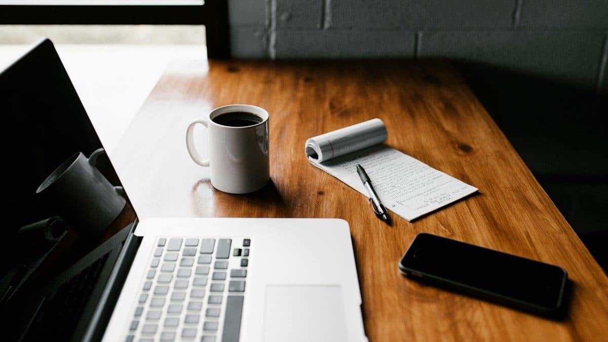 Portatile aperto su una scrivania di legno con tazza di caffè, blocco note, penna e smartphone